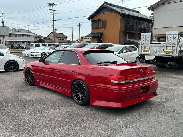 Modified red Toyota Mark II sedan with black wheels parked in a Japanese lot.